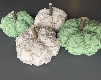 May include: Four decorative fabric pumpkins in shades of green and cream. Each pumpkin has a textured, looped design and a small, natural wooden stem. The pumpkins are arranged on a gray surface, perfect for fall decor.