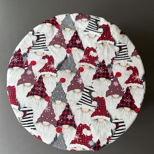 May include: Circular fabric item with a repeating pattern of Christmas gnomes. The gnomes have white beards, red and black plaid hats, and striped hats. The background is white.