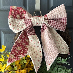 May include: A handmade wreath featuring a large, quilted bow with red floral and striped patterns. The bow is the focal point, complemented by a base of greenery, yellow forsythia, and peach-colored roses. The wreath hangs on a blue door.