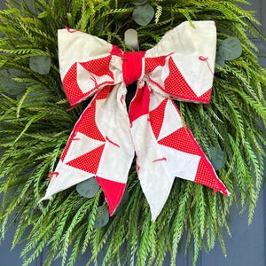 May include: A decorative bow crafted from red and white fabric with a polka dot pattern, featuring a central knot and two long, pointed tails. The bow is positioned over a lush, full wreath made of green fern-like foliage and round leaves.