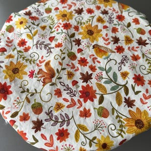 May include: A round fabric bowl cover featuring a white background with a colorful autumn-themed print. The pattern includes yellow sunflowers, orange and red flowers, brown acorns, green leaves, small red mushrooms, and a small brown squirrel.