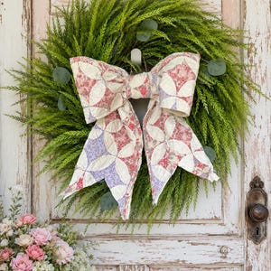 May include: A green wreath made of needle-like foliage and eucalyptus leaves, featuring a large, decorative bow crafted from quilted fabric. The fabric bow displays a patchwork pattern in shades of cream, muted red, and soft purple floral prints.