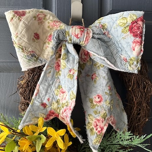 May include: A large, quilted fabric bow with a floral pattern of red roses and green leaves on a light blue background. The bow is attached to a grapevine wreath with yellow and green floral accents, creating a decorative piece for a door or wall.