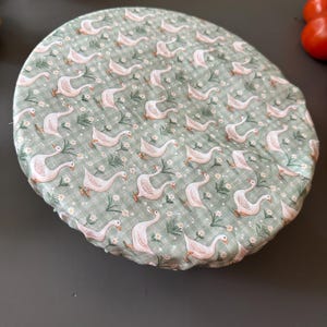 May include: A round fabric bowl cover featuring a repeating pattern of white geese and small white flowers on a sage green gingham background. The cover has an elasticized edge designed to fit snugly over the rim of a circular container.