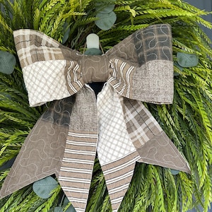 May include: A large decorative bow made of quilted fabric patches in shades of brown, beige, and white. The bow features various patterns including plaid, stripes, and cross-hatch designs with visible stitching, attached to a green leafy wreath.