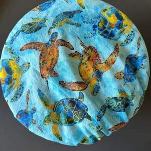 May include: A round, light blue fabric item featuring a repeating pattern of sea turtles in shades of blue, orange, and yellow. The turtles are depicted in various poses, creating a lively and aquatic theme.