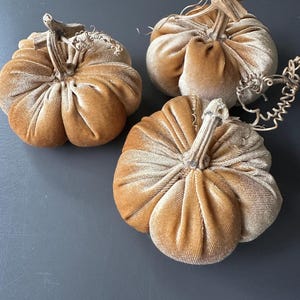 May include: Three plush, pumpkin-shaped decorations in varying sizes. The pumpkins are made of a soft, velvety fabric in a warm, golden-brown color. Each pumpkin has a realistic stem and is ideal for fall decor.