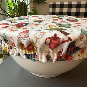 May include: A white ceramic bowl covered with a fabric bowl cover. The cover features a repeating pattern of garden gnomes, watering cans, and gardening tools in red, green, and yellow on a cream background. The cover has a ruffled edge.