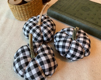 Könnte beinhalten: Drei schwarze und weiße Gingham-Kürbisse mit braunen Stielen. Die Kürbisse sind auf einer beigefarbenen Stoffoberfläche angeordnet.