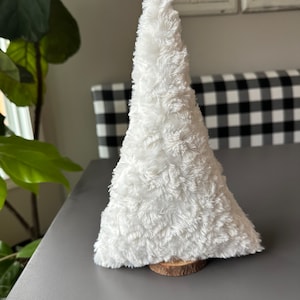 May include: A small, white, fluffy, decorative tree. The tree is cone-shaped and made of a soft, textured material. It is mounted on a small, round, wooden base. The tree is likely a holiday or seasonal decoration.