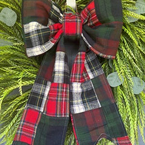 May include: A decorative bow made from a patchwork of plaid fabric featuring red, green, black, white, and yellow patterns. The bow has two large loops and two long, hanging tails with notched ends, all constructed from various tartan squares.