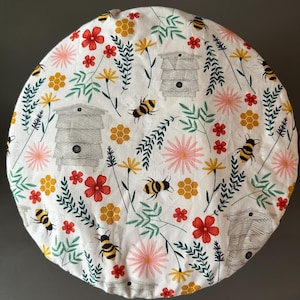May include: A round, white fabric item with a repeating pattern of bees, beehives, flowers, and honeycomb. The flowers are red, pink, and yellow, with green leaves. The bees are black and yellow. The beehives are gray.