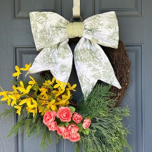 May include: A decorative wreath featuring a large white bow with a green toile pattern. The wreath is adorned with yellow forsythia, pink roses, and green foliage, creating a vibrant and welcoming display. The wreath is hanging on a gray door.