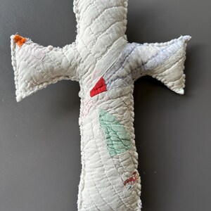 May include: A white quilted cross with a scalloped edge. The cross is made of patchwork fabric with different colors and patterns, including red, green, and blue.
