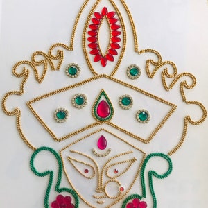 May include: A white background with a gold, green, and red beaded design of a Hindu goddess. The goddess has a red and gold crown, green earrings, and a red and gold face.