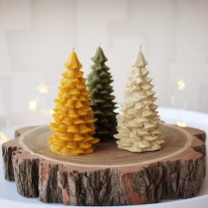 Puede incluir: Tres velas decorativas con forma de árbol en tonos amarillo, verde y crema. Las velas están colocadas sobre una rodaja de madera, que a su vez está sobre una bandeja blanca. Las velas tienen un diseño texturizado y en capas, que se asemeja a un árbol de Navidad. El fondo está borroso con luces de cadena.