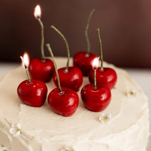 May include: A white iced cake topped with cherry-shaped candles, each with a lit flame. The cake is decorated with small white flowers, creating a celebratory dessert.