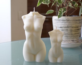 Op de afbeelding: Twee witte, gebeeldhouwde kaarsen in de vorm van een menselijk torso. De kaarsen staan op een glazen tafel met een plant in een pot erachter.