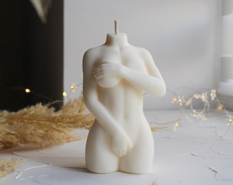 Op de afbeelding: Een witte kaars in de vorm van een menselijk torso. De kaars staat op een wit oppervlak.