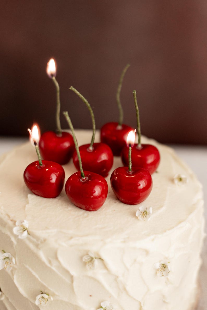 May include: A white iced cake topped with cherry-shaped candles, each with a lit flame. The cake is decorated with small white flowers, creating a celebratory dessert.
