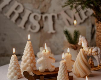 May include: A collection of lit, ivory-colored Christmas candles in various shapes, including Christmas trees, a gingerbread man, and a snowflake. A wooden sled is in the center. The word "CHRISTMAS" is in the background.