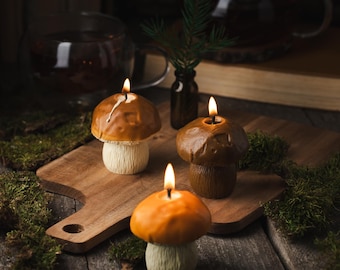 May include: Three mushroom-shaped candles on a wooden chopping board. The candles are lit and have a warm glow. The candles are surrounded by moss and a small glass bottle.