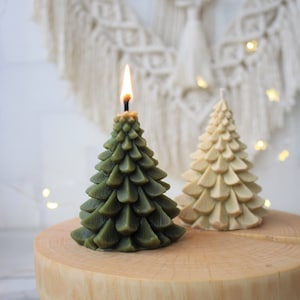 May include: Two Christmas tree shaped candles, one green and one cream, on a wooden surface. The green candle is lit.
