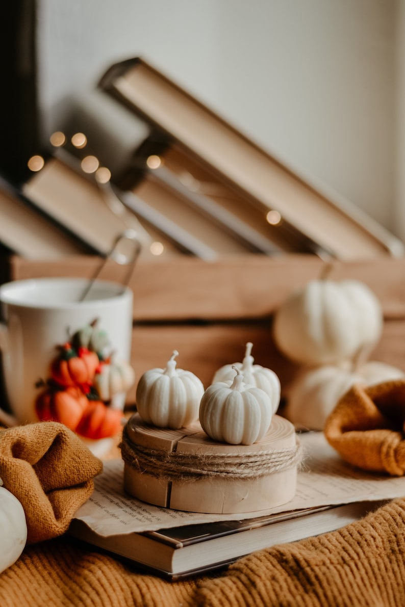 K&ouml;nnte beinhalten: Ein herbstliches Stillleben mit wei&szlig;en, k&uuml;rbisf&ouml;rmigen Kerzen auf einem Holzsockel, verziert mit Bindfaden. Eine Tasse mit K&uuml;rbisillustrationen, B&uuml;cher und ein warmfarbiger Pullover vervollst&auml;ndigen die Szene. Weiche Beleuchtung und ein verschwommener Hintergrund tragen zum saisonalen Ambiente bei.