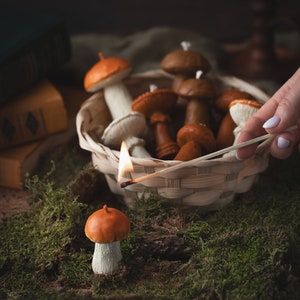 May include: A still life featuring mushroom-shaped candles in a wicker basket and on moss. The candles are various shades of brown, orange, and white. A lit match is held near the basket, and a single candle is in the foreground.