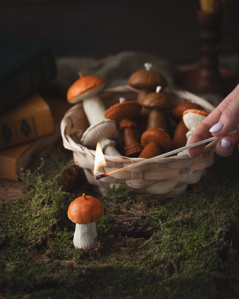 May include: A still life featuring mushroom-shaped candles in a wicker basket and on moss. The candles are various shades of brown, orange, and white. A lit match is held near the basket, and a single candle is in the foreground.