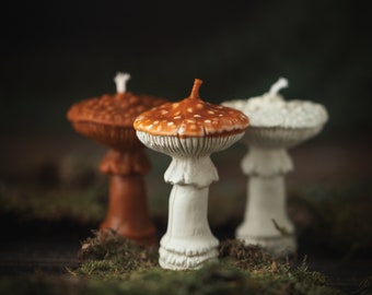May include: Three mushroom-shaped candles, two white with brown spots and one brown with white spots, are arranged on a bed of green moss.