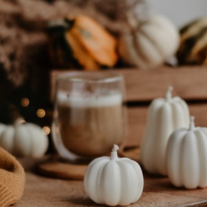 May include: A cozy autumn still life featuring white pumpkin-shaped candles on a wooden tray. A glass of coffee with foam sits behind the candles. Other pumpkins and gourds are in the background, with a yellow blanket.