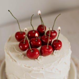 May include: A white iced cake decorated with cherry-shaped candles, some lit. The bright red cherries have green stems. Small white flowers are also on the cake, creating a celebratory dessert.