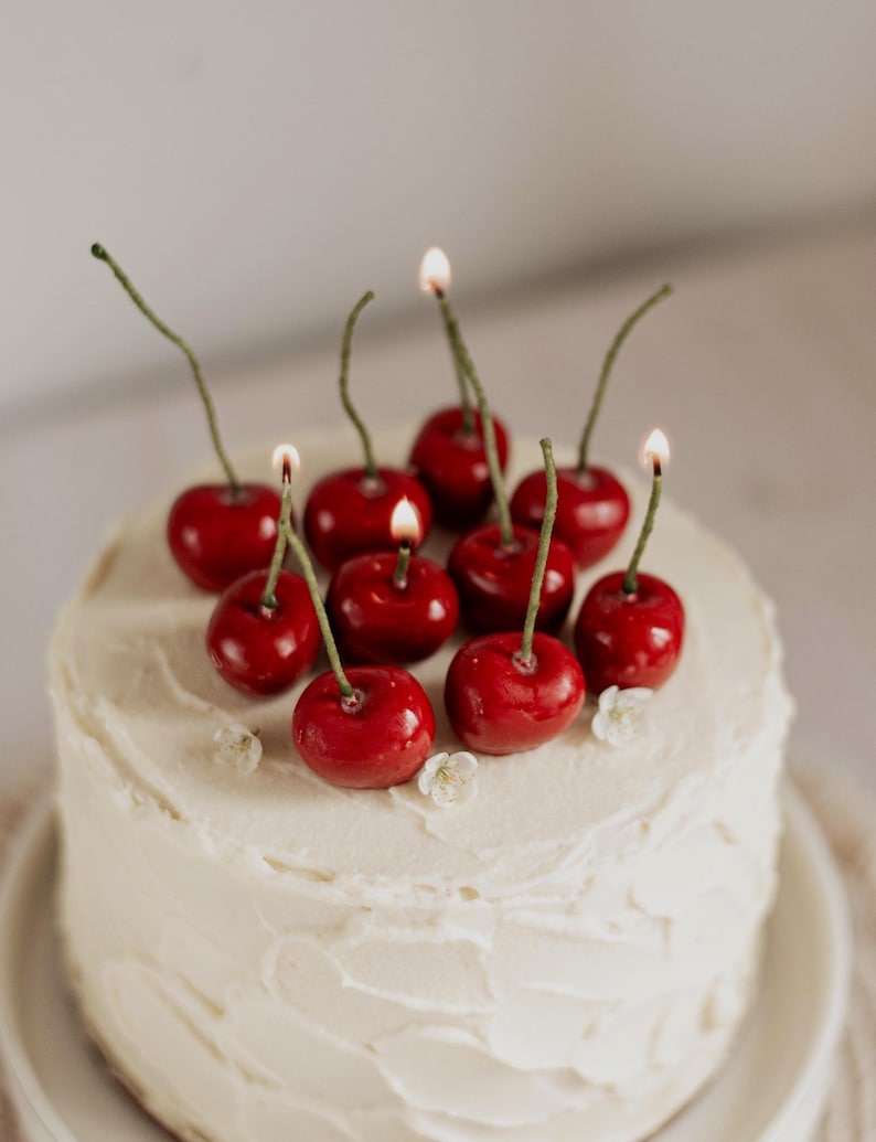 May include: A white iced cake decorated with cherry-shaped candles, some lit. The bright red cherries have green stems. Small white flowers are also on the cake, creating a celebratory dessert.
