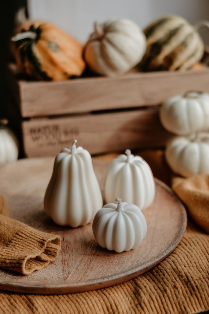 May include: A collection of white pumpkin-shaped candles on a wooden tray, with additional pumpkins in the background. The candles have a ribbed texture and wicks. The scene evokes a fall or harvest theme.
