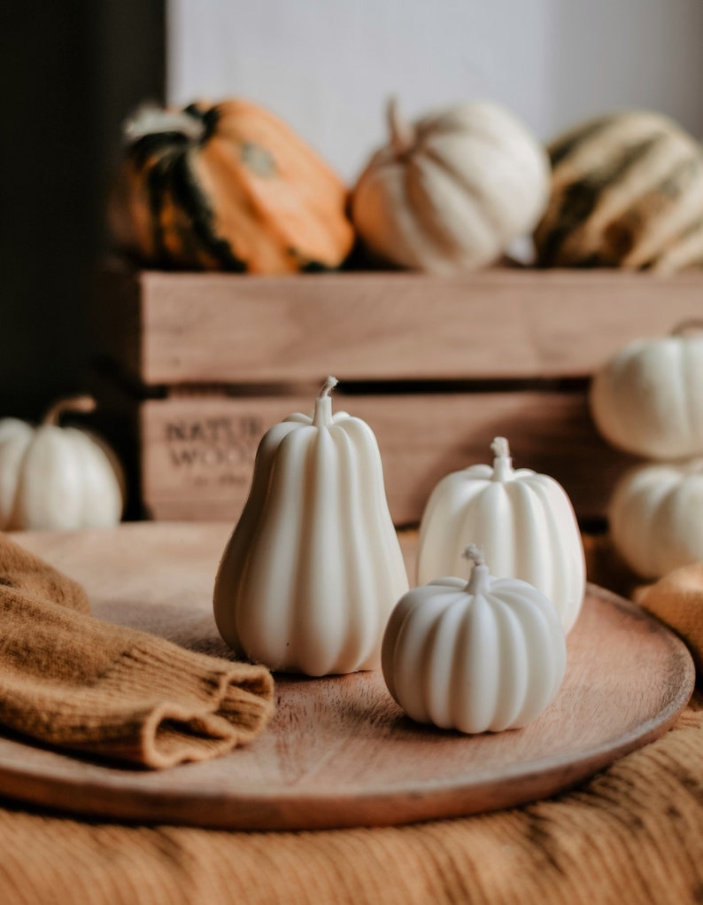 May include: A wooden tray displays several white pumpkin-shaped candles of varying sizes. Behind the candles, a wooden crate holds a collection of gourds in shades of orange, green, and white. A folded brown sweater rests on the tray.