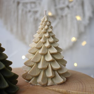 May include: Two Christmas tree candles, one is green and one is beige. The beige candle is on a wooden surface.