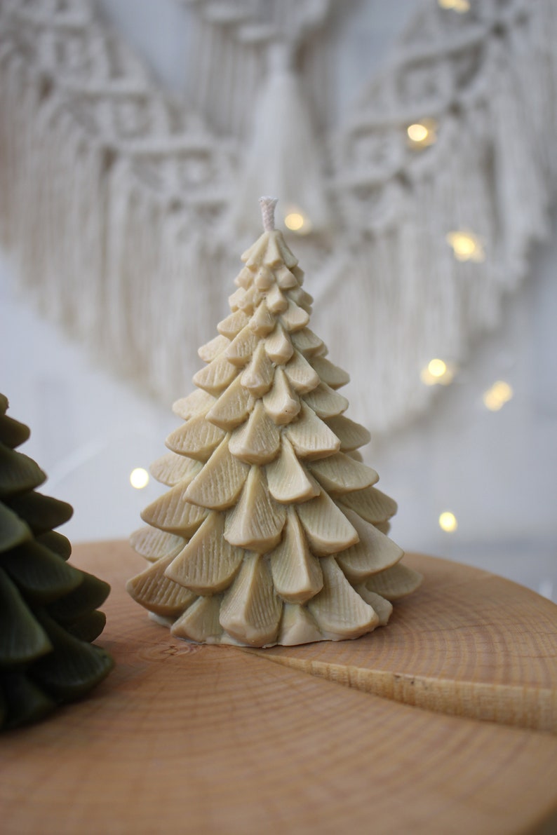 May include: Two Christmas tree candles, one is green and one is beige. The beige candle is on a wooden surface.