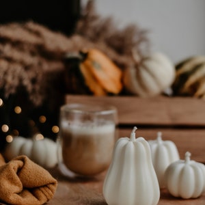 May include: A wooden tray holds white pumpkin-shaped candles, a glass of coffee, and a folded mustard-colored cloth. Other pumpkins and gourds are in the background, creating a fall harvest theme. The candles have a ribbed texture.