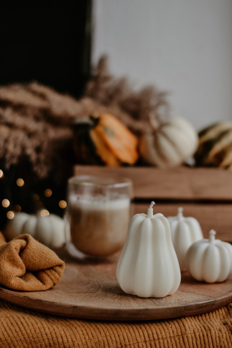 May include: A wooden tray holds white pumpkin-shaped candles, a glass of coffee, and a folded mustard-colored cloth. Other pumpkins and gourds are in the background, creating a fall harvest theme. The candles have a ribbed texture.