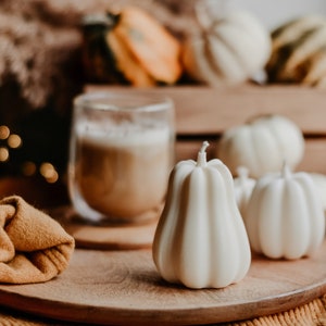 May include: A still-life arrangement featuring white pumpkin-shaped candles on a wooden tray. A glass of coffee and a folded mustard-colored cloth are also on the tray. Other pumpkins are in the background, creating a warm, autumnal theme.