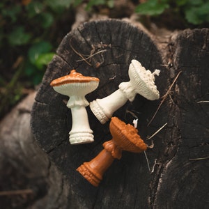May include: Three mushroom-shaped candles in cream, orange, and brown hues rest on a weathered tree stump. The candles have detailed caps and stems, with a natural, earthy aesthetic. The arrangement suggests a woodland or nature-inspired theme.