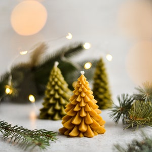 May include: Three yellow beeswax candles shaped like Christmas trees on a white surface with green pine branches.