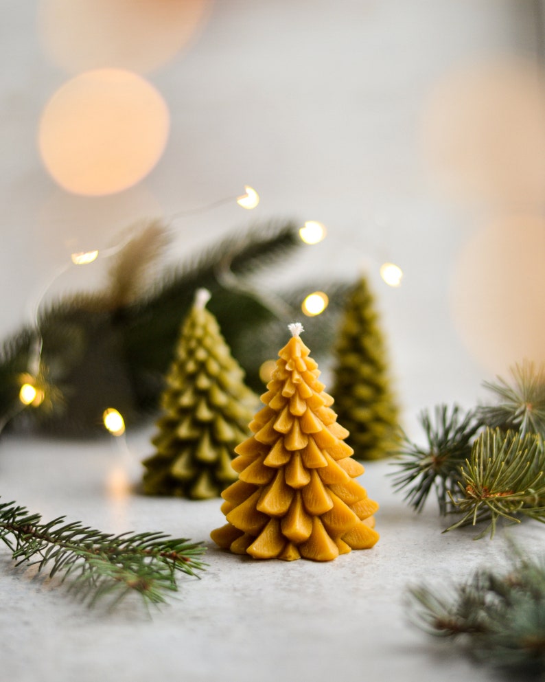 May include: Three yellow beeswax candles shaped like Christmas trees on a white surface with green pine branches.