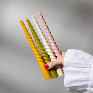Puede incluir: Una mano sostiene cinco velas en espiral en tonos amarillo, verde, blanco y rosa. Las velas tienen un diseño retorcido y no están encendidas. El fondo es gris liso.