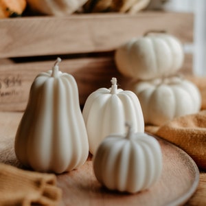 May include: A collection of white pumpkin-shaped candles of varying sizes, arranged on a wooden tray and a wooden crate. The candles have a ribbed texture and a wick at the top. Other pumpkins are in the background.
