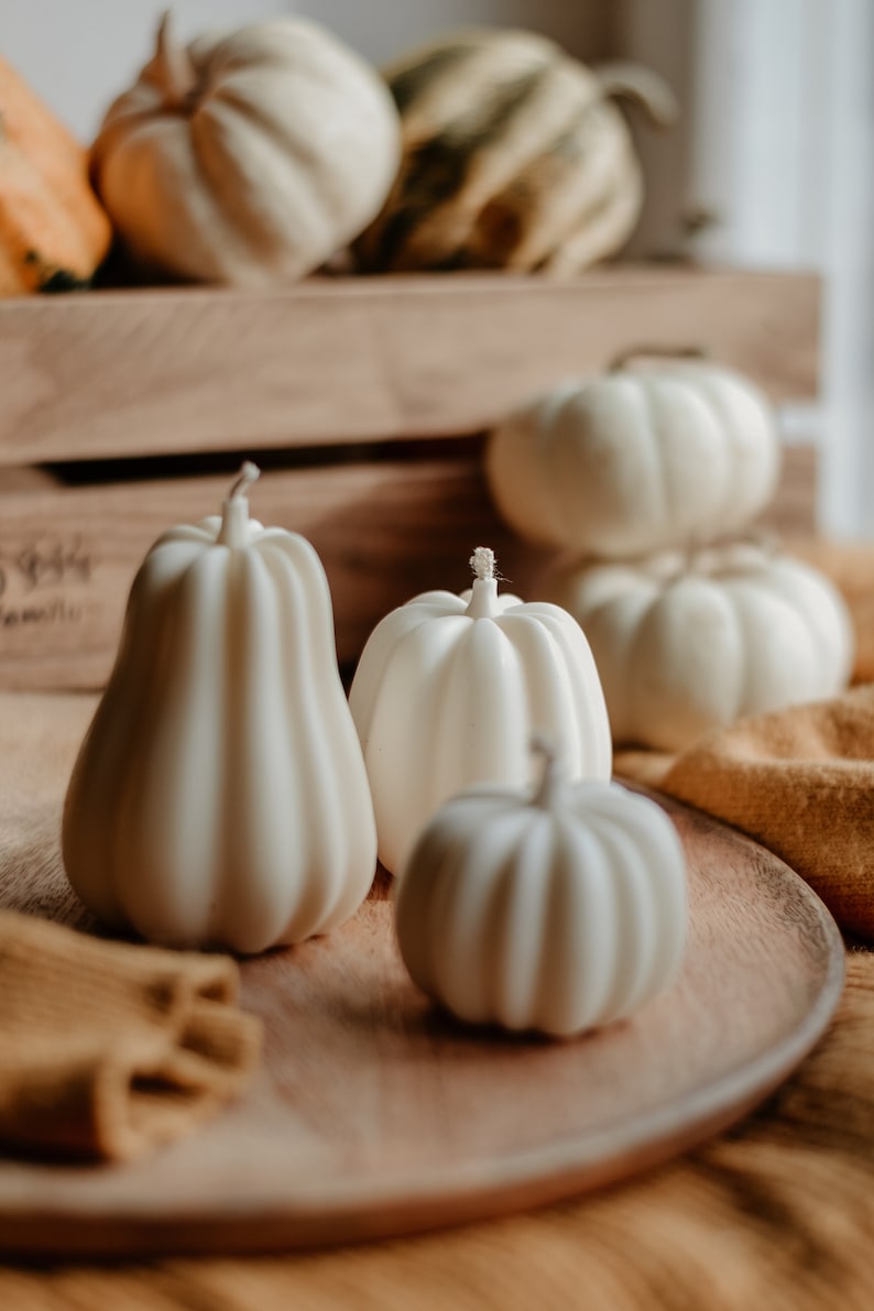 May include: A collection of white pumpkin-shaped candles of varying sizes, arranged on a wooden tray and a wooden crate. The candles have a ribbed texture and a wick at the top. Other pumpkins are in the background.