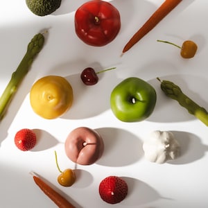 May include: A collection of fruit and vegetable shaped candles including an avocado, red apple, green apple, peach, garlic, strawberries, cherries, asparagus stalks, and carrots. Each item features a small white wick protruding from the top.