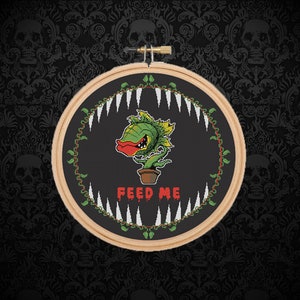 May include: A black and white cross-stitch embroidery hoop with a green, red, and yellow cartoon plant with teeth and the text "FEED ME".