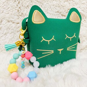 May include: A green cat-shaped purse with gold accents and a matching tassel keychain. The keychain also features a colourful beaded bracelet with the name "Amelia" spelled out.